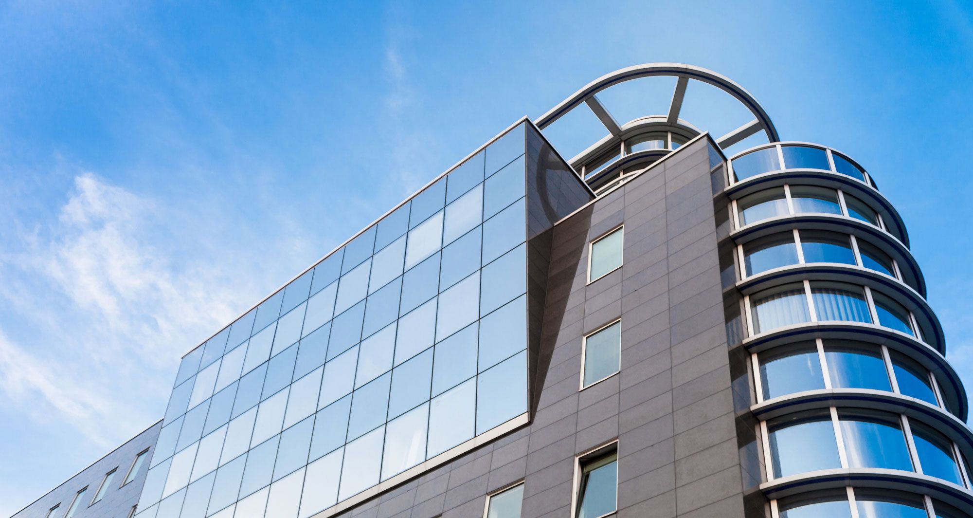 Modernes Bürogebäude mit großen spiegelnden Glasfenstern und einem geschwungenen Eckteil vor einem klaren blauen Himmel. Die architektonische Besonderheit aus Metall krönt das Dach.